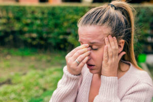 Woman experiencing a severe headache, holding her head and pinching the bridge of her nose in pain, outdoors.