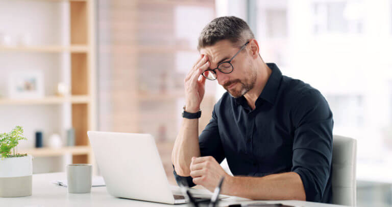An office worker sits at his laptop, wincing in discomfort as he experiences a chronic headache or migraine.
